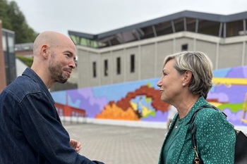 Cécile Neven souriante, serre la main d'un représentant de la Dinantaise. En arrière plan, une immense fresque aux couleurs vives.