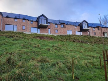 Vue d’une rangée de nouveaux logements sociaux mitoyens en briques rouges, construits sur une colline herbeuse. Les maisons ont des toits en ardoise noire, des baies vitrées modernes et, pour certaines, un petit balcon.